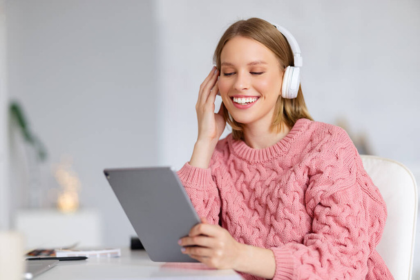 Счастливая молодая женщина в трикотажном свитере улыбается и слушает музыку в наушниках, сидя за столом - Фото, изображение