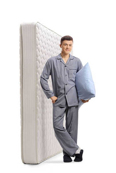 Full length portrait of a young man in pajamas leaning on a bed mattress and holding a pillow isolated on white background - Zdjęcie, obraz