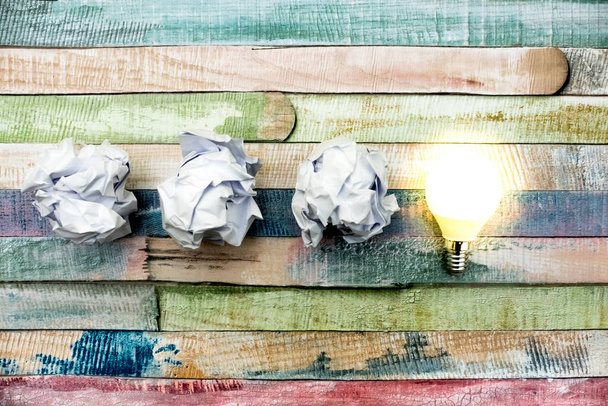 Nieuw ideeënconcept met verfrommeld wit papier en gloeiende gloeilamp op kleurrijke houten tafel. - Foto, afbeelding