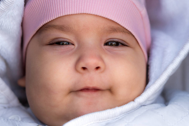 concept bébé, enfance, émotion - gros plan de mignon portrait souriant visage de nouveau-né potelé aux yeux bruns bébé édenté sans dents 6-9 mois regardant la caméra allongée dans une veste blanche chapeau rose dans une poussette - Photo, image