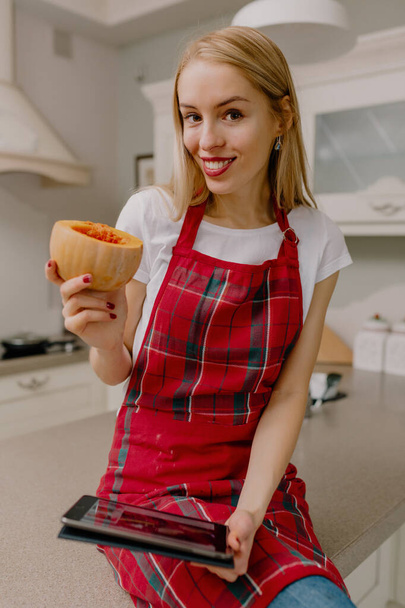 Attraktive Frau mit langen blonden Haaren und Schürze kocht in der Küche mit Tablette. Hochwertiges Foto - Foto, Bild