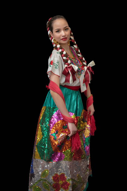 Mulher latina vestida como uma poblana de porcelana com uma saia bordada em lantejoulas coloridas, um xale vermelho, arcos trançados e um fundo preto sorridente - Foto, Imagem