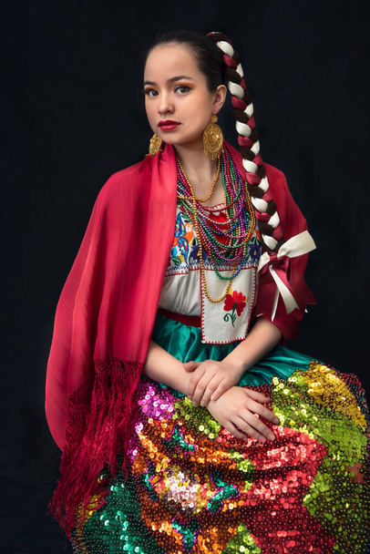 Latinská žena oblečená jako porcelánová poblana se sukní vyšívanou v barevných flitrech, červeným šátkem, splétanými luky a usměvavým černým pozadím - Fotografie, Obrázek