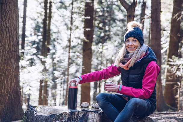 Een glimlachende vrouwelijke toerist neemt een pauze voor warme thee uit een thermoskan en een maaltijd op het bergpad. - Foto, afbeelding