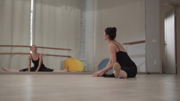 Voor een repetitie in een danszaal strekt een prachtige ballerina haar armen en benen uit terwijl ze op de vloer zit op een spleet voor een spiegel - Video