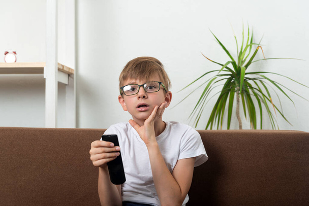 A szemüveges fiú távirányítóval a kezében TV-t néz. A gyerek a 18-as csatornát nézi+. - Fotó, kép