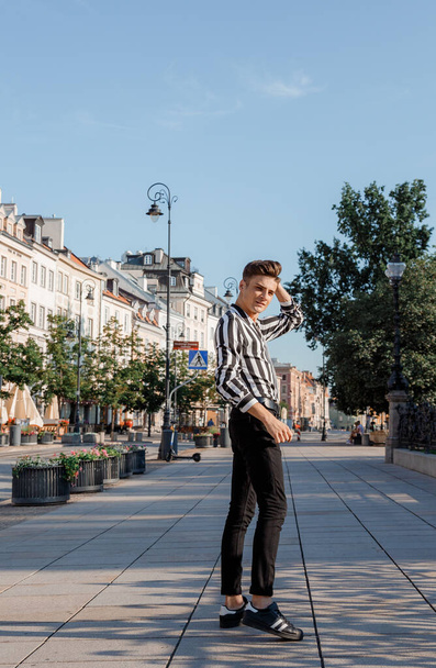 A young man walks on the sidewalk. Guy model posing Stylish guy in black and white shirt and black pants. City walk. Travel. Poland. Warsaw. - Фото, изображение
