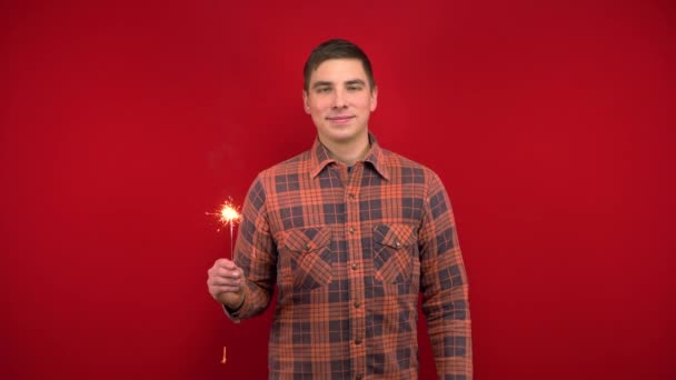 Ein junger Mann im roten Hemd hält eine Wunderkerze in der Hand. Dreharbeiten im Studio auf rotem Hintergrund. - Filmmaterial, Video