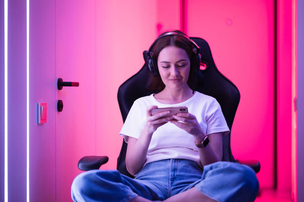Cybersport-Gamer spielt Handyspiel auf dem Smartphone sitzend auf einem Gaming-Stuhl im neonfarbenen Raum. - Foto, Bild