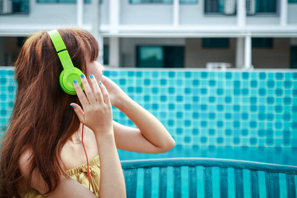 Una hermosa chica sentada en una silla al lado de la piscina, llevaba un auricular musical. - Foto, imagen