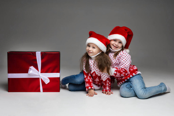 Прекрасні маленькі дівчата в капелюхах з різдвяним подарунком. великий різдвяний подарунок у червоному папері та білому банті в студії на сірому фоні
. - Фото, зображення