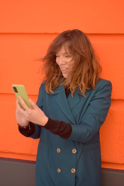 Mujer joven sonríe amablemente a lo que ve en su teléfono de carcasa verde. fondo de la pared naranja y ella está usando un abrigo de color verde azulado - Foto, Imagen