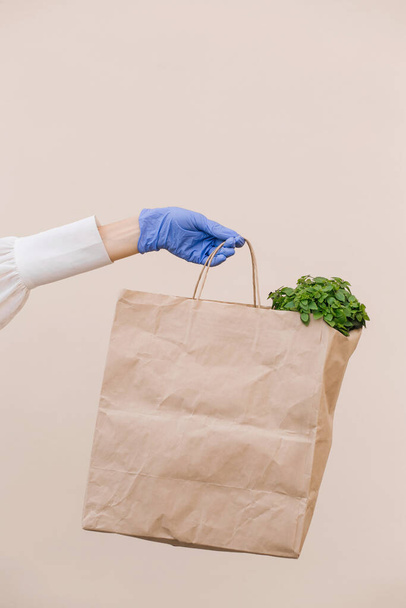 Female hands in blue gloves holds a brown paper bag with products, vegetables, isolated over beige background. Safe food delivery to your home.Quarantine, coronavirus.Copy space. - Φωτογραφία, εικόνα