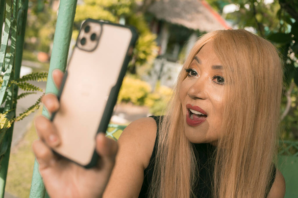 Svobodná Asiatka středního věku s obarvenými blond vlasy pózuje pro selfie. Cítit se mladá, sexy a sebevědomá. Venkovní nastavení. - Fotografie, Obrázek