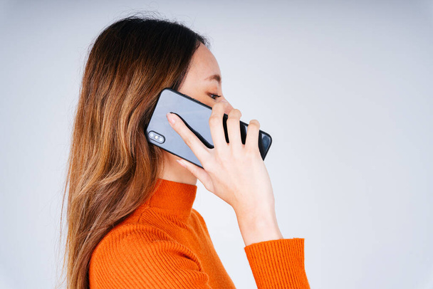 Boční pohled - Asijky v oranžovém svetru mluví na telefonu izolovat na bílém pozadí. - Fotografie, Obrázek