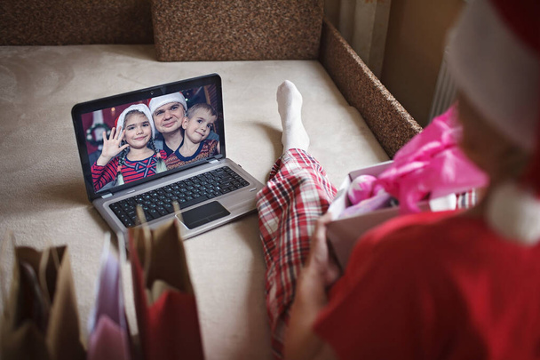 Sichere Online-Weihnachtsfeier. Seniorin mit Weihnachtsmannmütze feiert mit ihrer Familie virtuell mit Internet und Notizbuch. Videoanruf. Bleiben Sie zu Hause, Urlaub in der Ferne, Blick auf den Bildschirm, Lebensstil - Foto, Bild