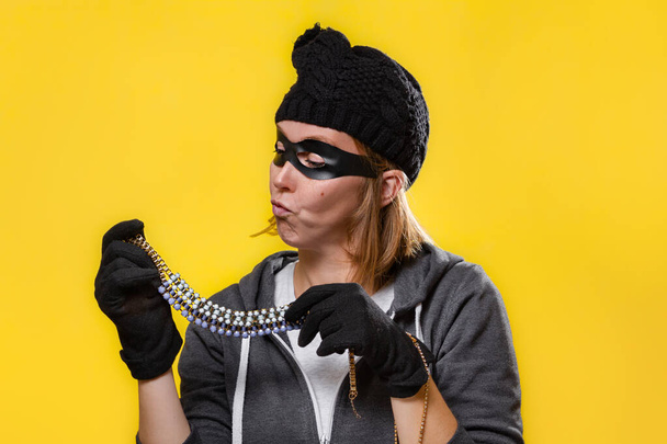 Поняття крадіжки. Жінка в капелюсі, масці і рукавичках тримає коштовності в обох руках і оглядає їх. Жовтий фон і простір для копіювання
. - Фото, зображення