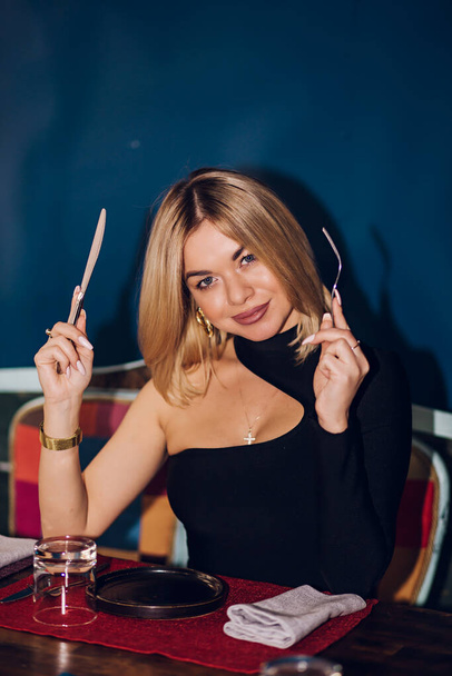 A woman holds a fork, knife at the table in a restaurant. Blonde girl wearing a black dress and sitting in a cafe, close-up portrait. Food, drinks, romantic evening concept - Photo, Image