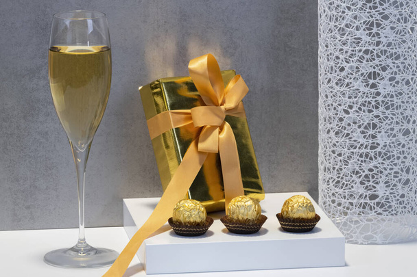 Selective focus of a golden wrapped gift box with wrapped chocolate sweets and a glass of champagne. Celebration and luxury concept. - Photo, Image
