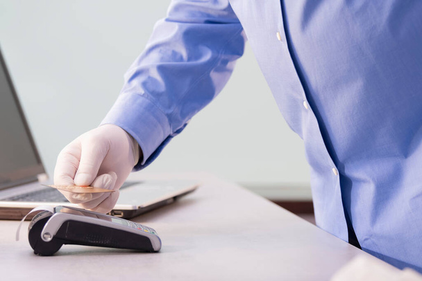 Close-up de um homem com a mão em uma luva segurando um cartão de crédito sobre um leitor de cartão de crédito em um fundo fora de foco. Conceito de empresa e prevenção. - Foto, Imagem
