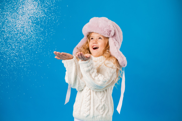 linda niña en rosa sombrero peludo jugando con nieve falsa sobre fondo azul en el estudio - Foto, Imagen