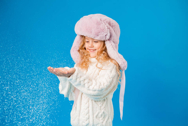 linda niña en rosa sombrero peludo jugando con nieve falsa sobre fondo azul en el estudio - Foto, Imagen