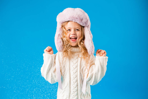 mignonne petite fille en chapeau de fourrure rose jouant avec de la fausse neige sur fond bleu en studio - Photo, image