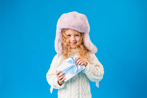 Cute smiling blonde girl in pink hat and white knitted sweater holding gift - Photo, image
