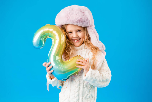 little girl in winter clothes smiling and holding a balloon in shape of number two on blue background - Foto, imagen