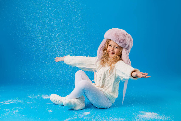 cute little girl in pink furry hat playing with fake snow on blue background in studio - Foto, afbeelding