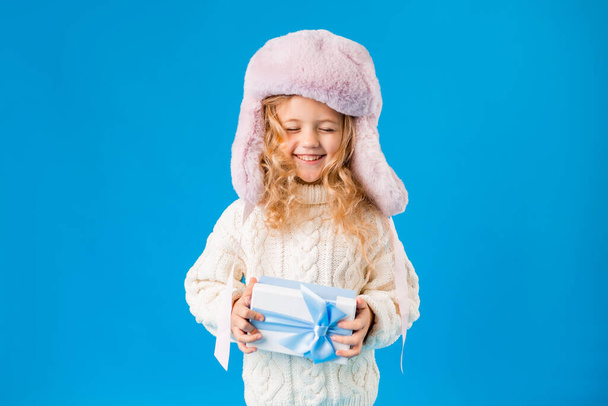 Cute smiling blonde girl in pink hat and white knitted sweater holding gift - Fotó, kép