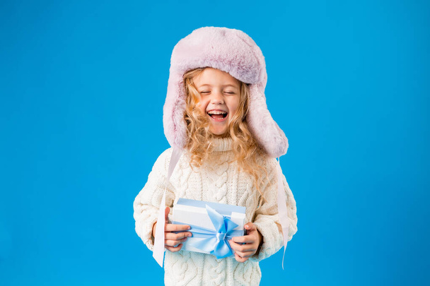 Cute laughing blonde girl in pink hat and white knitted sweater holding gift - Foto, Imagem