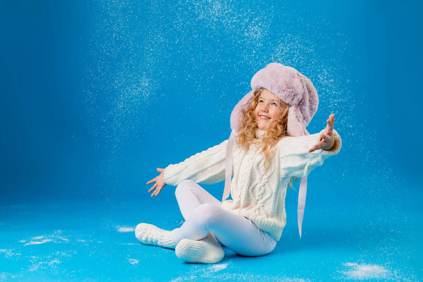 cute little girl in pink furry hat playing with fake snow on blue background in studio - Foto, Imagem