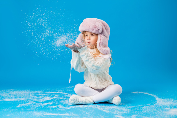 cute little girl in pink furry hat playing with fake snow on blue background in studio - Photo, Image