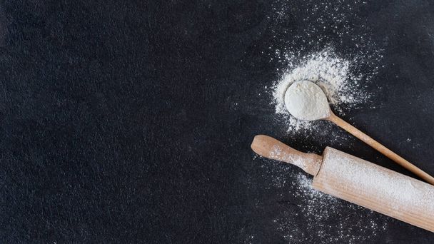 Wheat flour in a wooden spoon with rolling pin over black surface top view. Baking background for your design. - Foto, Imagen