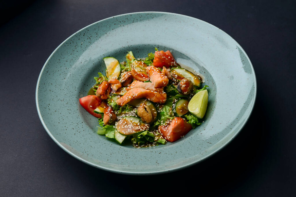 Salad with fish. Fresh vegetable salad with salmon fish fillet and sesame seeds on top served in a blue plate over black background. Asian cuisine concept. - Fotografie, Obrázek