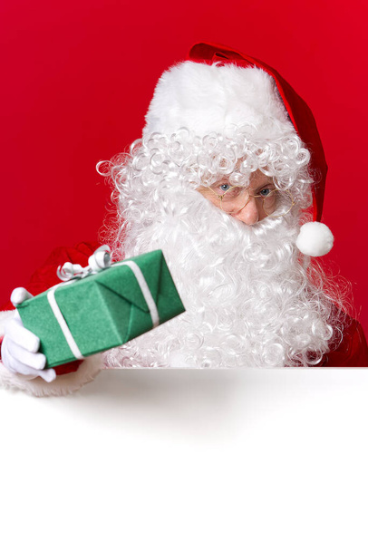 Joulupukki pitää vihreää lahjapakkausta kädessä seisoessaan tyhjän valkoisen laudan takana. kopioi tila. - Valokuva, kuva