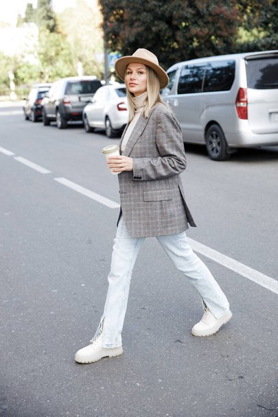 A Woman in a grey jacket and hat crosses the road with cars. A blonde girl holds a glass of coffee. Street style - Foto, Imagem