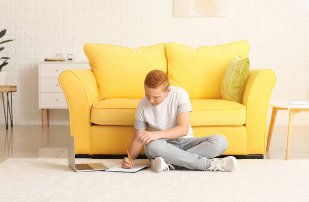 Мальчик-подросток, выполняющий домашнюю работу, сидя на полу дома - Фото, изображение