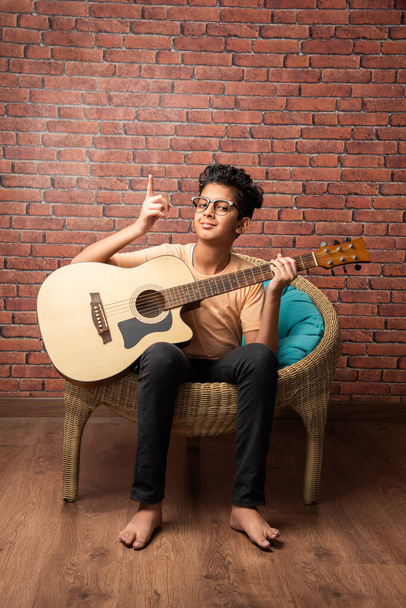 Indian asian boy playing acaustic guitar while sitting against white background or brick wala on chair - Foto, immagini