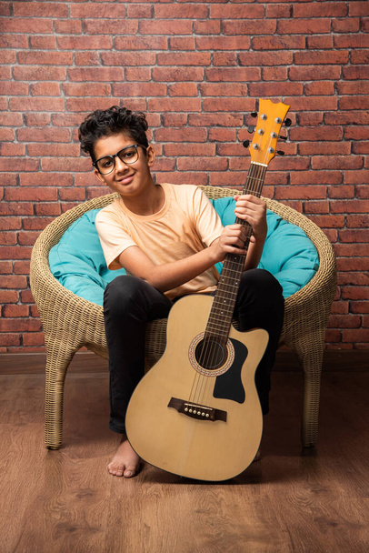 Indian asian boy playing acaustic guitar while sitting against white background or brick wala on chair - Foto, Imagem