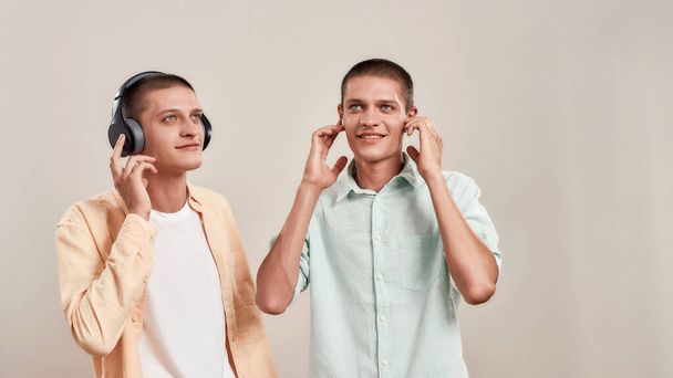 Enjoying favourite playlist. Two young caucasian twin brothers in casual wear listening music in headphones and wireless earphones - Fotoğraf, Görsel