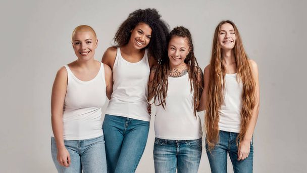 Portrait of four young diverse women wearing white shirts and denim jeans smiling at camera while posing together isolated over grey background - Фото, изображение