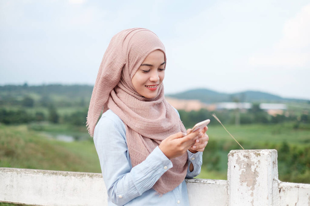 農場の牛の白い柵の近くのスマートフォンを見ている女性イスラム教徒の獣医師。ヘルスケアとテクノロジーの概念. - 写真・画像