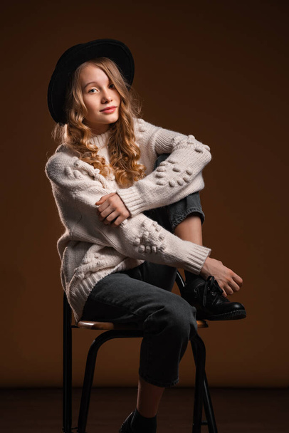 Charming blonde teen girl in black hat posing in studio on brown background. Concept of beauty and elegance - Foto, imagen