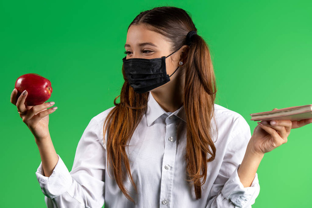 A girl in a mask, in a school white shirt, an apple in one hand and a book in the other. Green background. High quality photo - Photo, Image