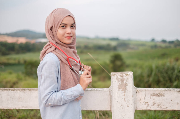The beautiful young female veterinarian wearing hijab with stethoscope stands near the white fence in the farm cow - Photo, Image