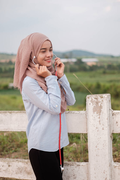 The beautiful young female veterinarian wearing hijab with stethoscope stands near the white fence in the farm cow - Photo, Image