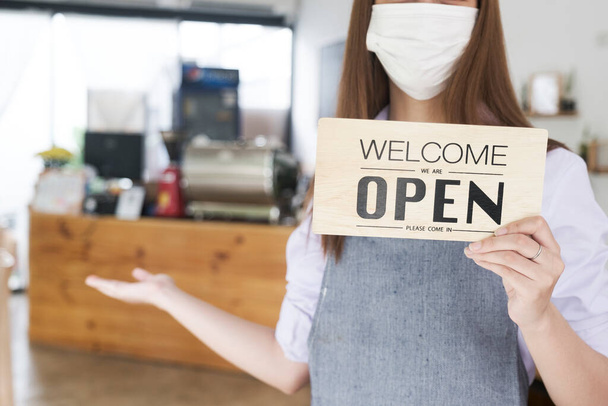 Business owner open restaurant as new normal. Happy coffee cafe owner with protective face mask holding open sign while standing at cafe doorway. - Photo, Image