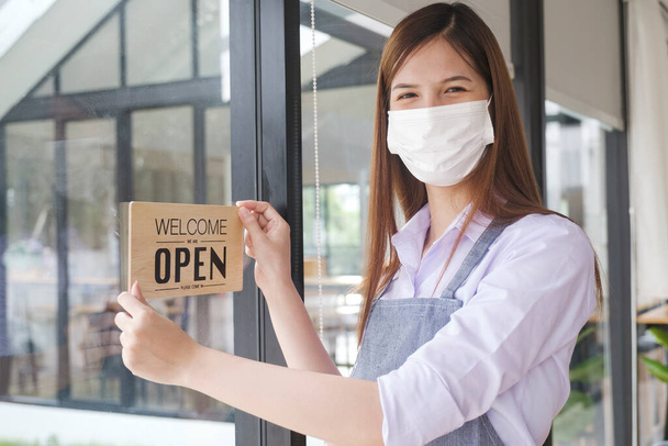 Business owner open restaurant as new normal. Happy coffee cafe owner with protective face mask holding open sign while standing at cafe doorway. - Foto, imagen